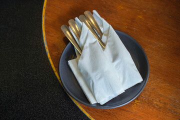 Black plate with spoon, fork and tissue on restaurant wooden table