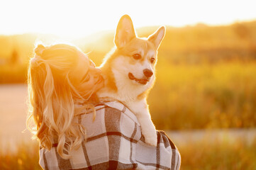 Woman holding Pembroke Welsh Corgi dog during golden summer sunset, hugging tenderly, sharing joy happy moment. Concept of caring for a pet, a walk in nature.