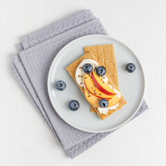 Top view of homemade sweet sandwich made with crispbread decorated with cream cheese, sliced nectarine fruit, blueberries and flax seeds served on plate on textile napkin on white wooden background