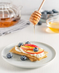 Sweet sandwich made with crispbread decorated with cream cheese, sliced nectarine fruit, blueberries and flax seeds served on plate with honey pouring from wooden dipper on table with glass tea pot
