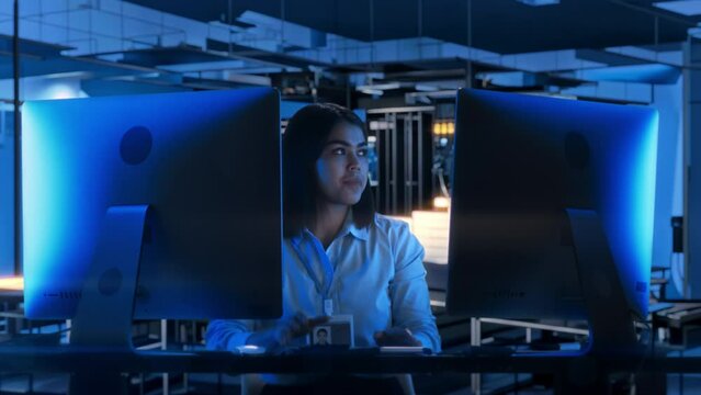 Young female IT specialist sitting at desk with dual monitor and working on computer in dark data center with blue neon light