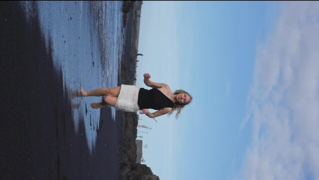 Woman wearing a black T-shirt and white skirt enjoys a peaceful day at the beach, running in the water and admiring the ocean. She runs on the sand, happy and carefree. Slow motion. Vertical view