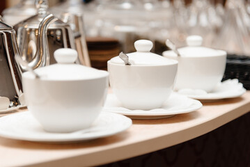 three small white tea cups with lids and spoons on saucers on counter, tableware for cafes, restaurant equipment