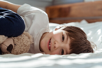 Portrait of little happy toddler boy lying on bed with his teddy bear plush toy looking at camera smiling at sunny morning