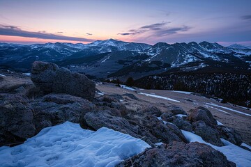 The setting sun paints the sky with twilight hues above a mountain range, as snow patches and rugged rocks add texture to the scene.