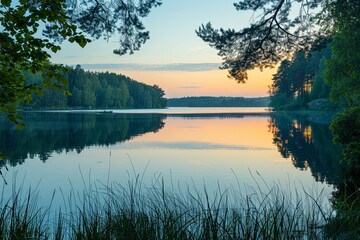 Fototapeta premium As twilight descends, the gentle hues of sunset reflect upon a calm lake, flanked by silhouetted trees and a peaceful sky.