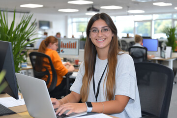 A young woman with glasses sits at her office workstation, surrounded by the vibrant atmosphere of a busy professional environment, while working on a laptop.Beginning of professional life, first job.