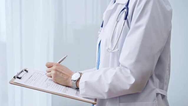 Female doctor taking notes on clipboard. Close-up of a unrecognizable female healthcare professional writing and filling um medication history records form while standing straight in clinic