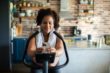 Young pregnant latina woman exercising on an indoor bike and using a smartphone