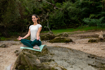 In the early morning hours, a stunning girl in sportswear sits on a large rock by the river beach in a meditation pose.