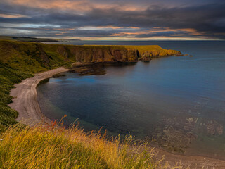 Sunset casts a warm glow over a sandy cove between rugged cliffs of Scotland