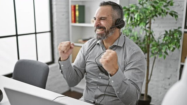 A mature man with a beard and headset celebrates in an office, showcasing joy in a professional setting.