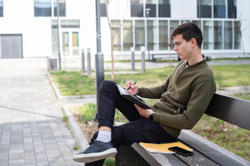 A young male student holding a notebook and sitting on a bench in front of a building, taking notes and learning for the exam on a sunny day