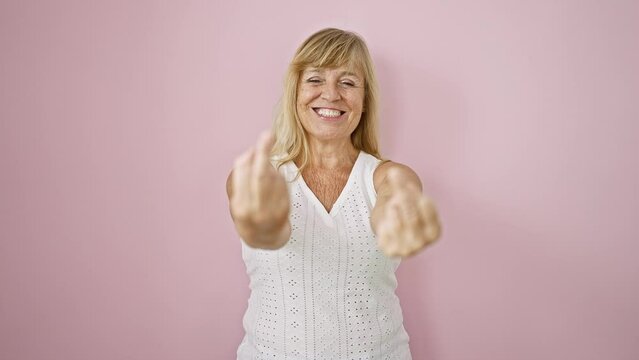 Cheerful middle age blonde woman flashes a confident smile while cheekily flipping the bird, standing against an isolated pink background