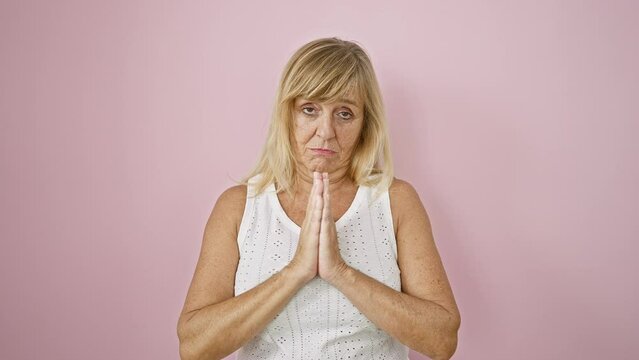 Beautiful mature blonde woman exudes serious expression, urgently clasping hands together in please gesture, standing isolated over vivid pink background