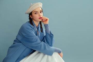 Fashionable confident woman wearing trendy spring outfit with white beret, blue coat, posing on blue background. Studio portrait. Copy, empty, blank space for text