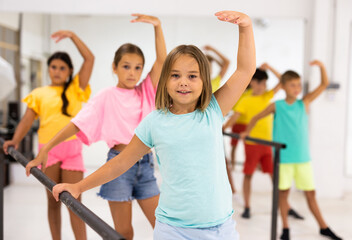 Portrait of happy preteen girl warming up near ballet bar during group class in dance school.
