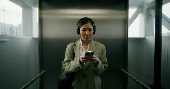 A woman in formal clothes and headphones uses a mobile phone while taking the elevator down to the railway station platform