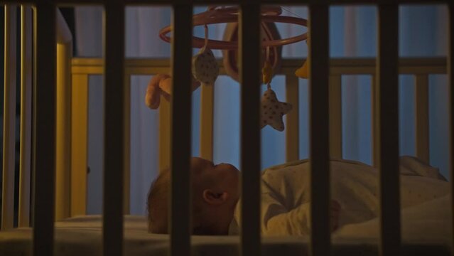 Small child lies in a crib at night getting ready for sleep. The moonlight coming through the window paints the atmosphere of deep night. A night lamp illuminates a small baby in a crib