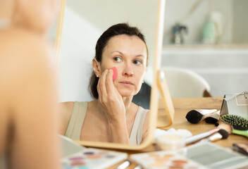 Calm middle-aged woman applying make-up foundation on her face by means of sponge sitting in front of the mirror