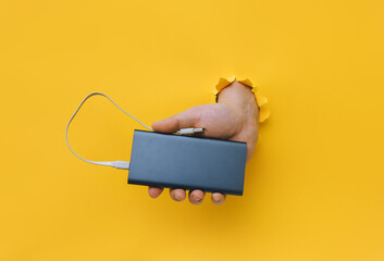 A man's hand with a gray metal power bank protrudes from a torn hole in yellow paper. Concept of...