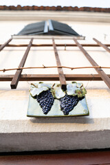 A rectangular ceramic plaque with a grape pattern is displayed on a fence with vinegrapes on a...