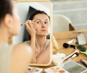 Focused adult woman meticulously shaping eyebrows with brush in front of tabletop mirror among...