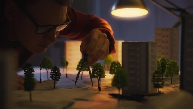 A white male, European, equipped with a tool, is thoughtfully designing a landscape under the light of a table lamp. At night, an architect undertakes the development of a residential area