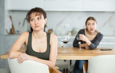 Offended middle-aged woman sitting in the kitchen while other woman talking angrily to her - obrazy, fototapety, plakaty