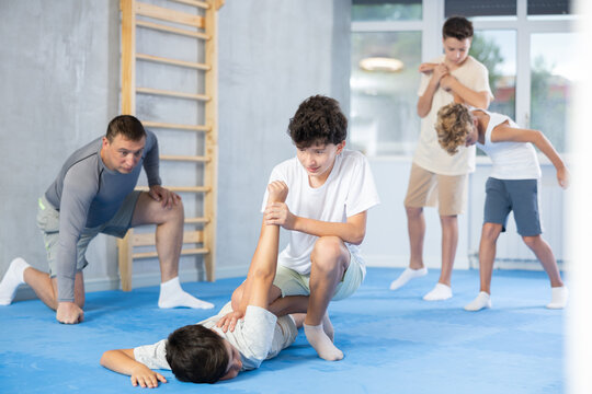 Concentrated boy learning effective self defence techniques in sparring, practicing painful pronating wristlock on male opponent lying face down on floor at modern sports studio