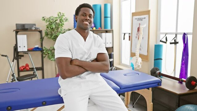 A confident young man in a white medical uniform stands with arms crossed in a brightly lit physical therapy clinic.