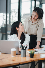 Two professional women engage in a collaborative discussion at a well-organized office desk, with one sitting and the other standing, sharing a moment of friendly interaction.