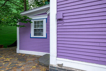 The exterior of a vibrant purple colored wooden wall is covered in horizontal clapboard siding. The building has white trim with thin blue lines. The green maple tree overhangs the sloping roof. 