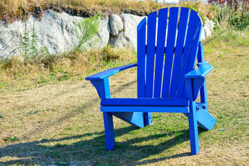 A vibrant empty royal blue color Adirondack chair in a lush green grassy garden. There's a rock or...