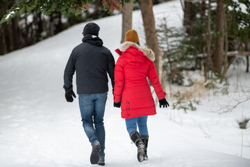 A senior man wearing a black winter jacket with jeans and an older female with a warm red coat, brown hat, jeans and winter boots walk outdoors on a trail covered with fresh snow in the woods. 