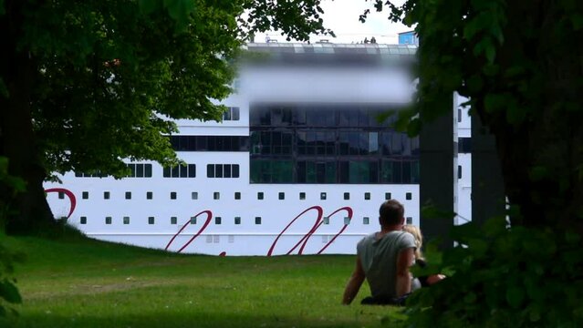 A Tranquil Scene Capturing A Couple Resting On Grass, Gazing At A Massive Cruise Ship Passing Behind A Veil Of Greenery - Stockholm, Sweden