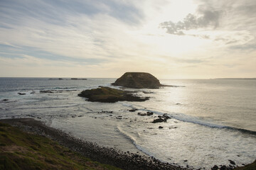 Coastal landscape with a small rocky island surrounded by ocean waves under a cloudy sky at sunset.