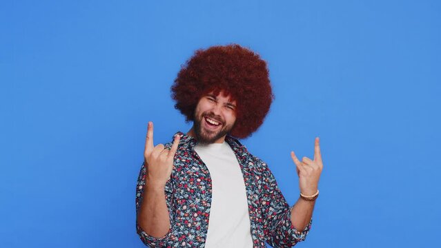 Overjoyed happy Caucasian man showing rock n roll gesture by hands, cool sign shouting yeah with crazy expression dancing rejoicing victory win success. Stylish young guy isolated on blue background