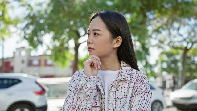 A thoughtful young asian woman stands outdoors in a city park, displaying casual fashion and natural beauty.