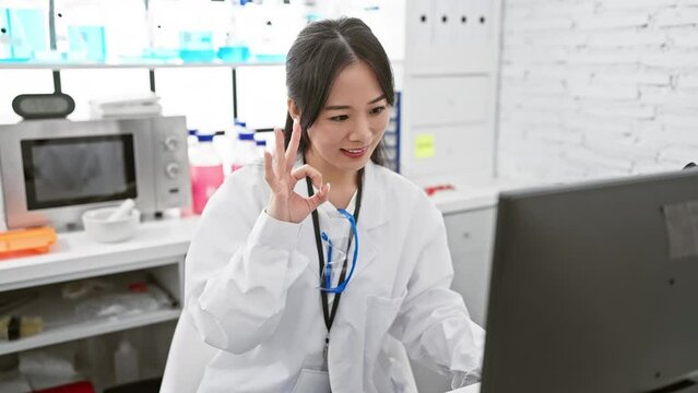 Confident young chinese scientist woman, radiant with success, flashes an 'ok' sign, flashing fingers in her pristine white lab coat. big smiles paint a positive expression on her face in the lab.