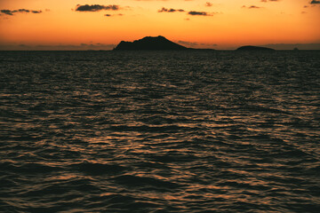 Islands rising on the horizon under an orange sky reflected by the gentle sea at sunset