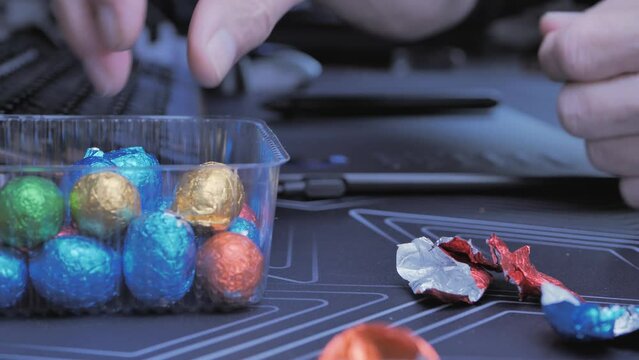 Adult man eating chocolate Easter eggs while working on computer at home, focus on hands taking colorful snacks