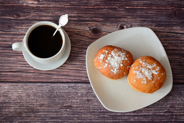 A cup of black coffee with a spoon on a saucer and a plate with two donuts in powdered sugar on a wooden table.