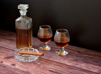 A crystal decanter and two glasses of cognac on a dark wooden table, next to a Cuban cigar in a glass ashtray.