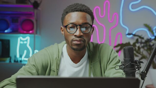 Gen Z African American blogger doing post production of digital media on laptop when sitting at table with mic in studio with multicolored neon decorations