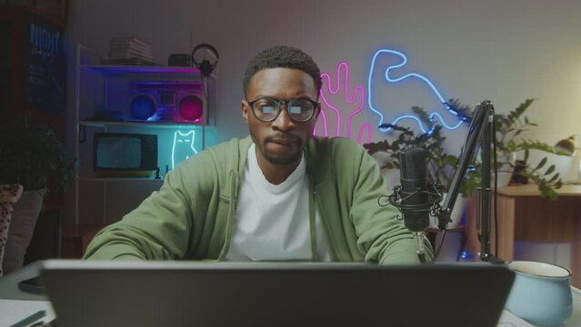Young Black blogger sitting at desk with mic in video studio with glowing neon decorations, looking at laptop screen and browsing the Internet, when working on social media content