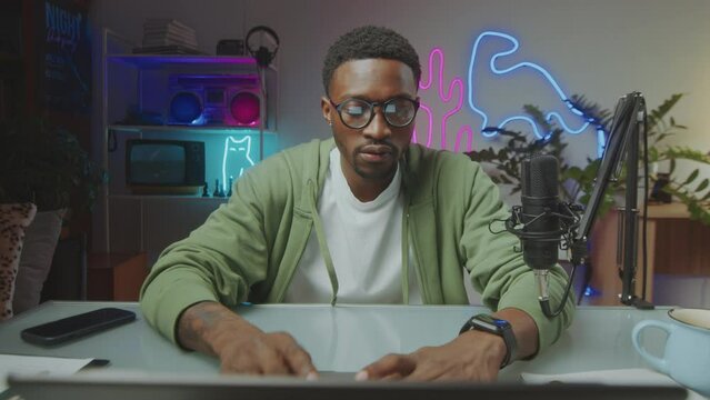 Young Black blogger opening laptop and typing on keyboard, doing post production or browsing the Internet at desk in modern video studio with neon light. Zoom shot