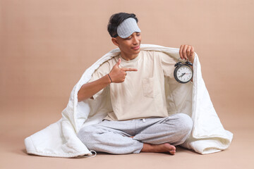 Young Asian man with sleep mask, sitting on floor covered with blanket and pointing at alarm clock