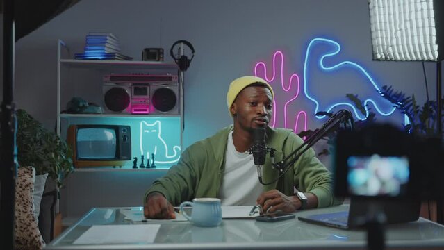 Young Black video blogger sitting in trendy studio with neon decorations and talking in mic in front of digital camera, going live or filming content for social media