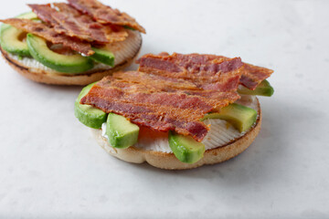 A view of an open faced bagel featuring bacon strips, avocado slices and cream cheese.